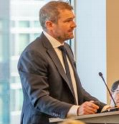 Christian Leathley speaking at a podium. Wearing a grey suit and a shite shirt and grey tie. He has fair skin and short cut grey hair.