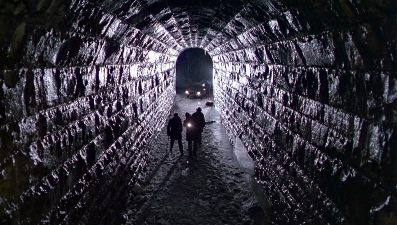 Dark tunnel with a car at the end with lights on, There are three people in the tunnel with a torch.