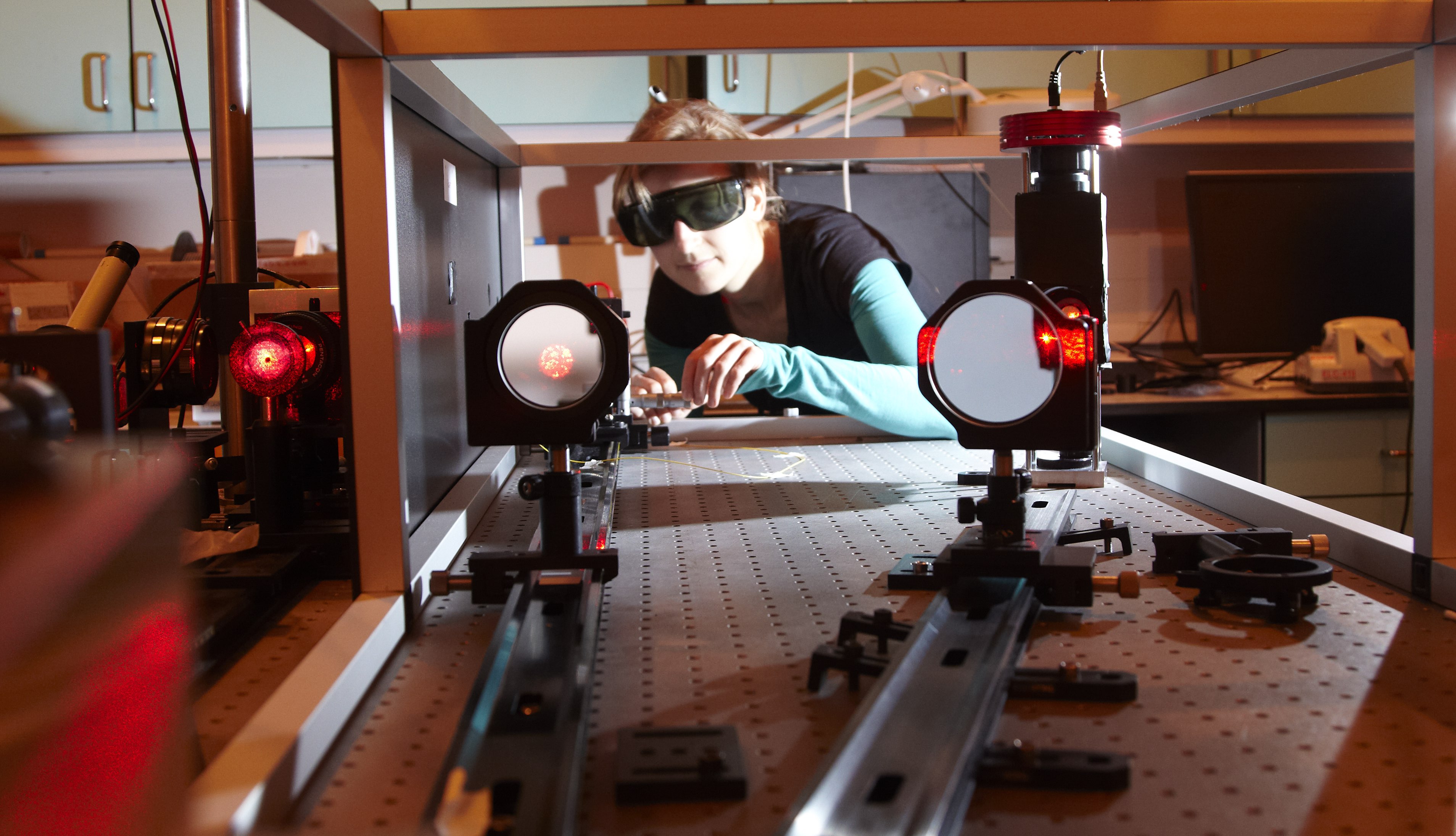 Woman wearing dark protective glasses looks through at a laser-lit optics bench