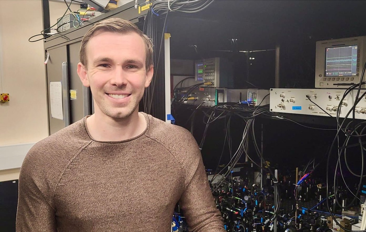 Photo of Dr Alex Guttridge smiling at camera next to an optics bench