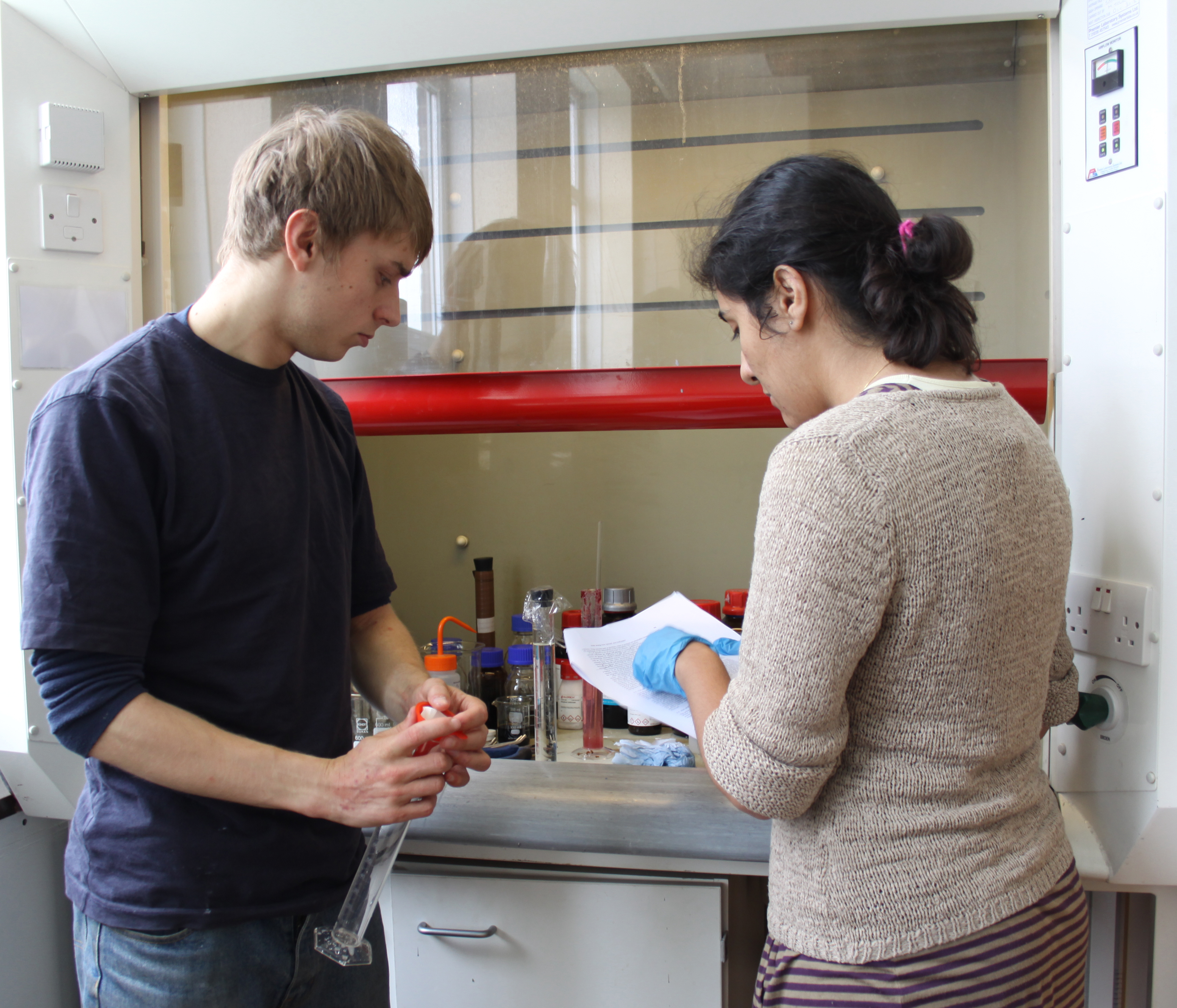 Male and female student checking notes by fume cupboard. One holds a test tube in his hands.