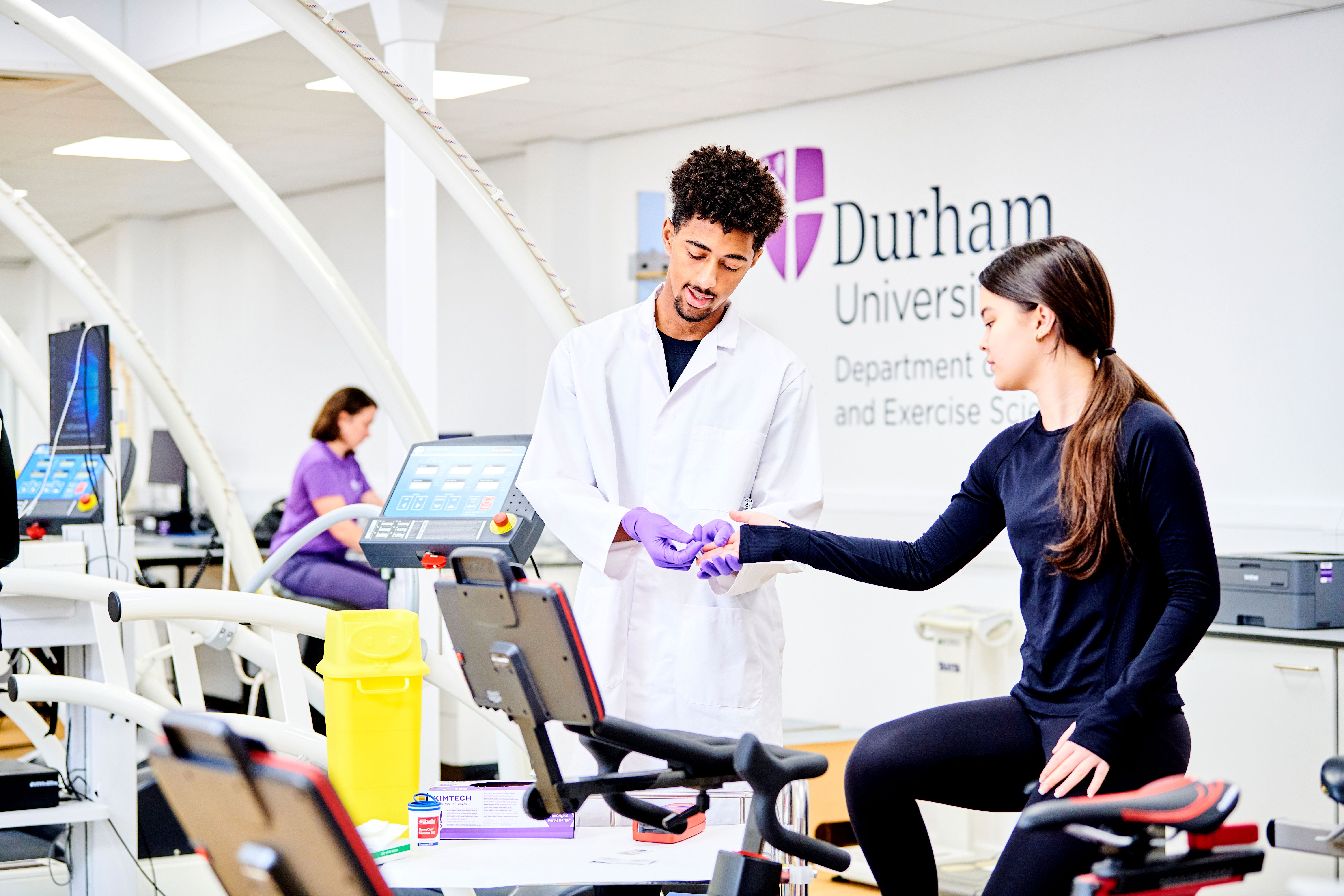 Male student in lab coat taking a pin prick blood sample from a female student sitting on a bench