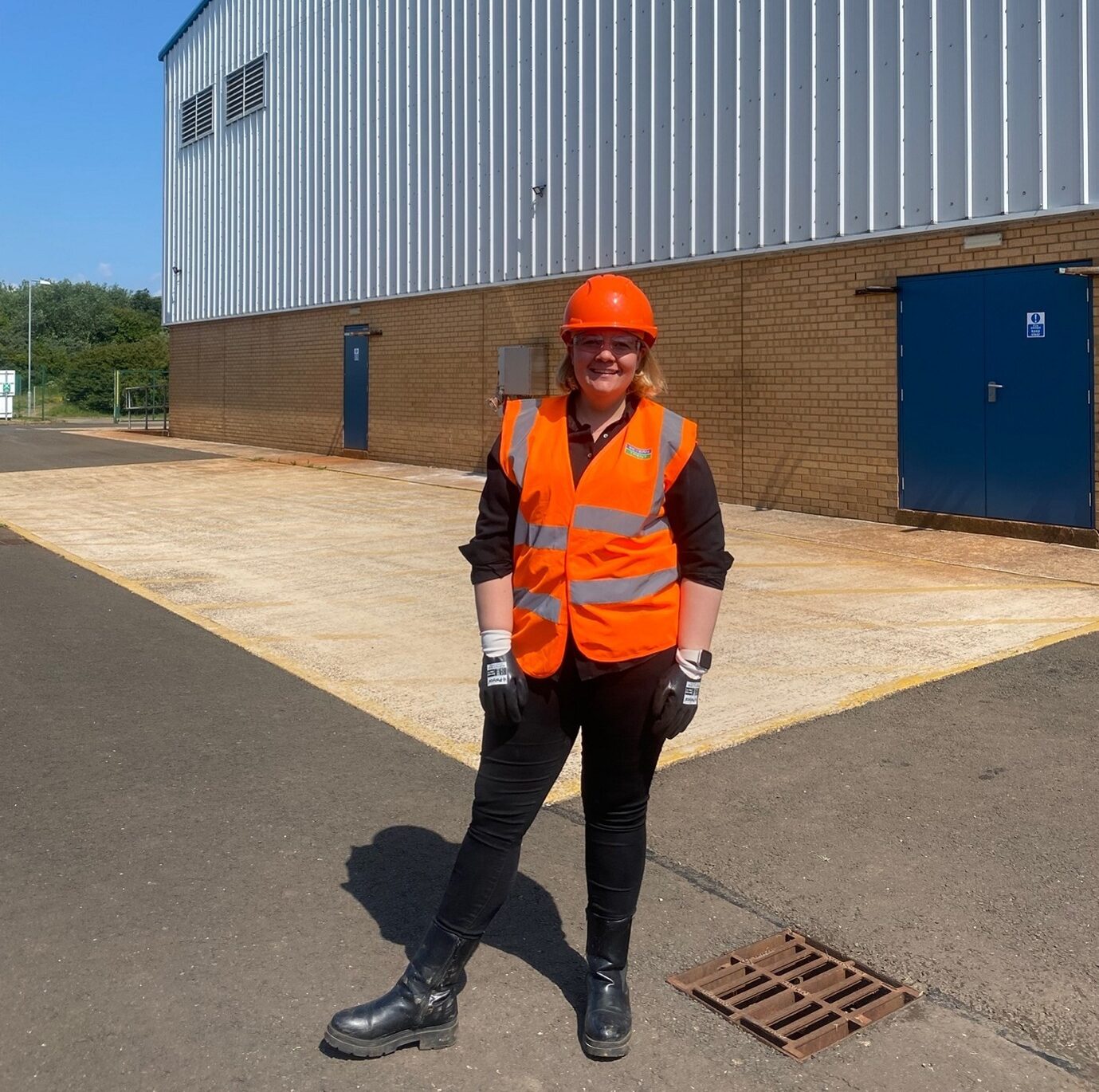 A PhD student in an orange hi-vis vest and hard hat outside an industrial building