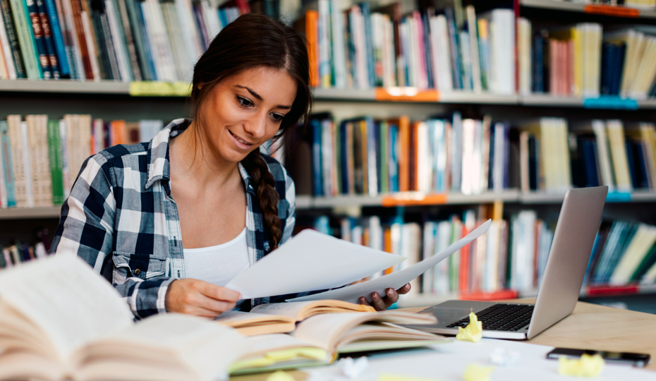 Student making notes using books and a laptop in front of bookshelves