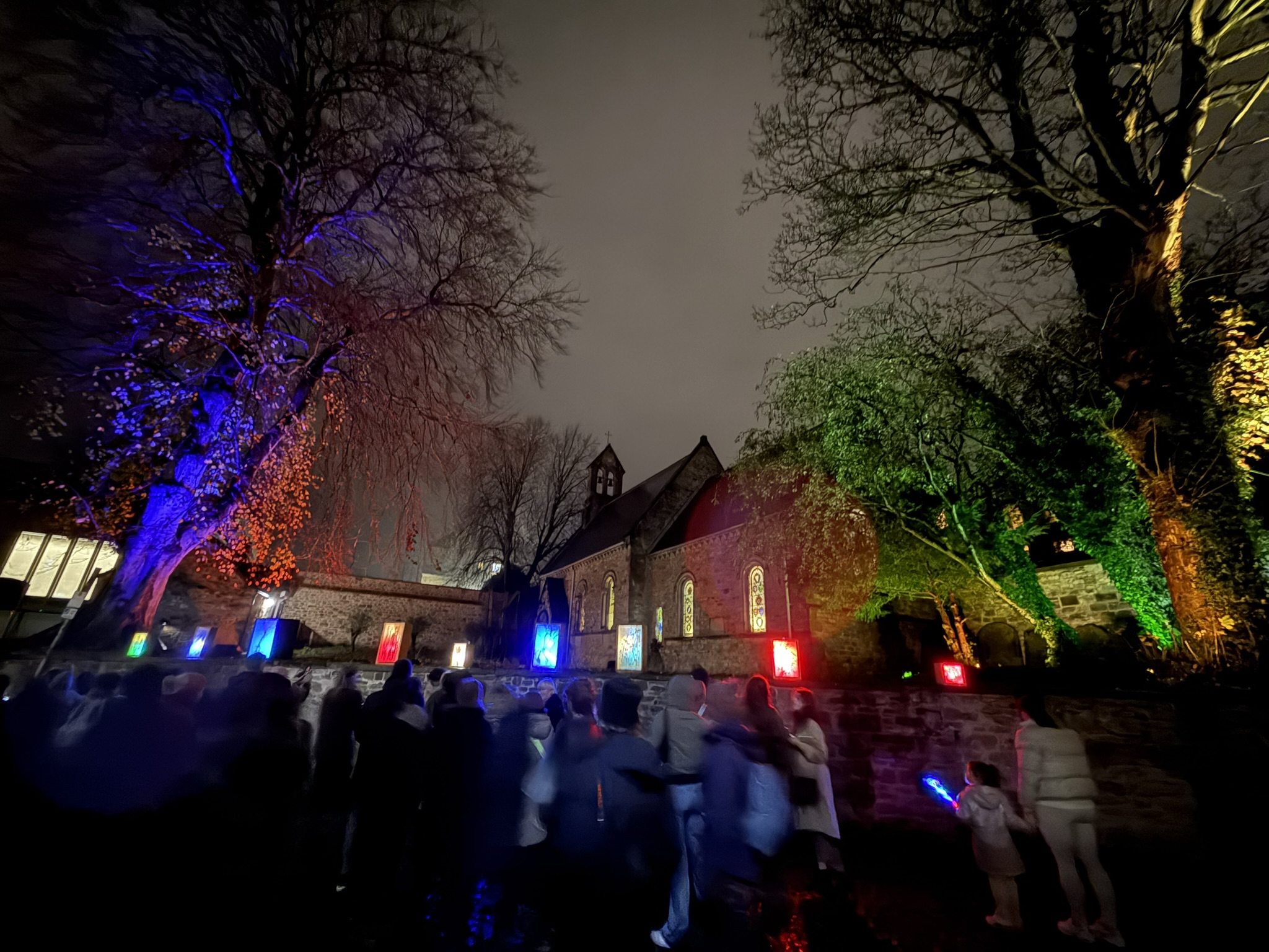 An old building with trees at either side lit up in different colours. On a wall in front of the building are illuminated boxes and portraits.