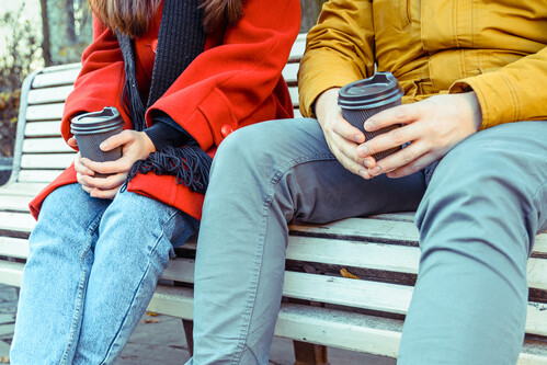 Close up of two students sitting on a bench, holding coffee cups in their hands