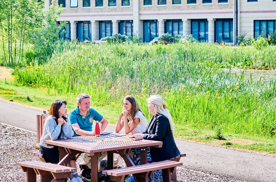 Staff sit at a wooden table chatting and relaxing. It's a sunny day and they are dressed in casual summer clothes. A pond surrounded by green plants and bushes is visible behind them. The Mountjoy buildings can be glimpsed in the background