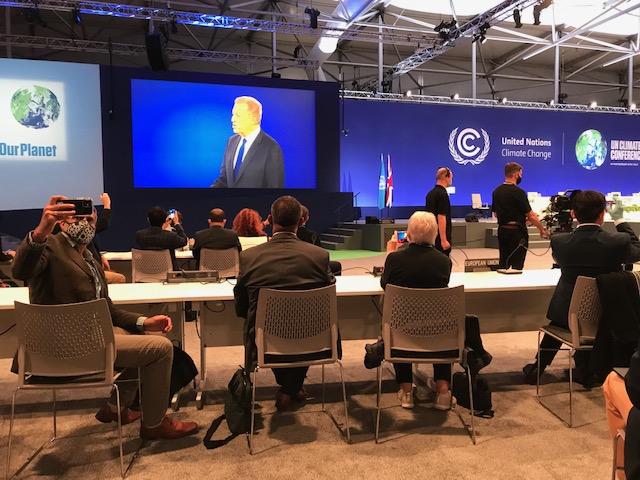 Two people sitting with their backs to the camera, looking at a speaker on a stage