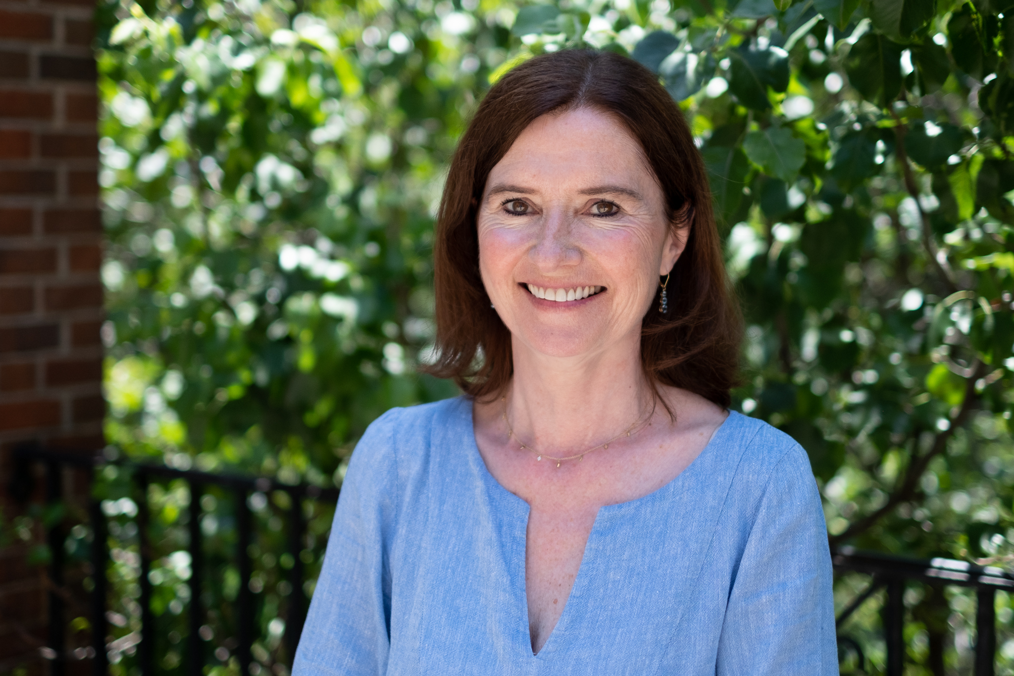 Prof Gráinne de Burca (NYU) smiling big. They are wearing a light blue cardigan and is standing in front of a wall of greenery.