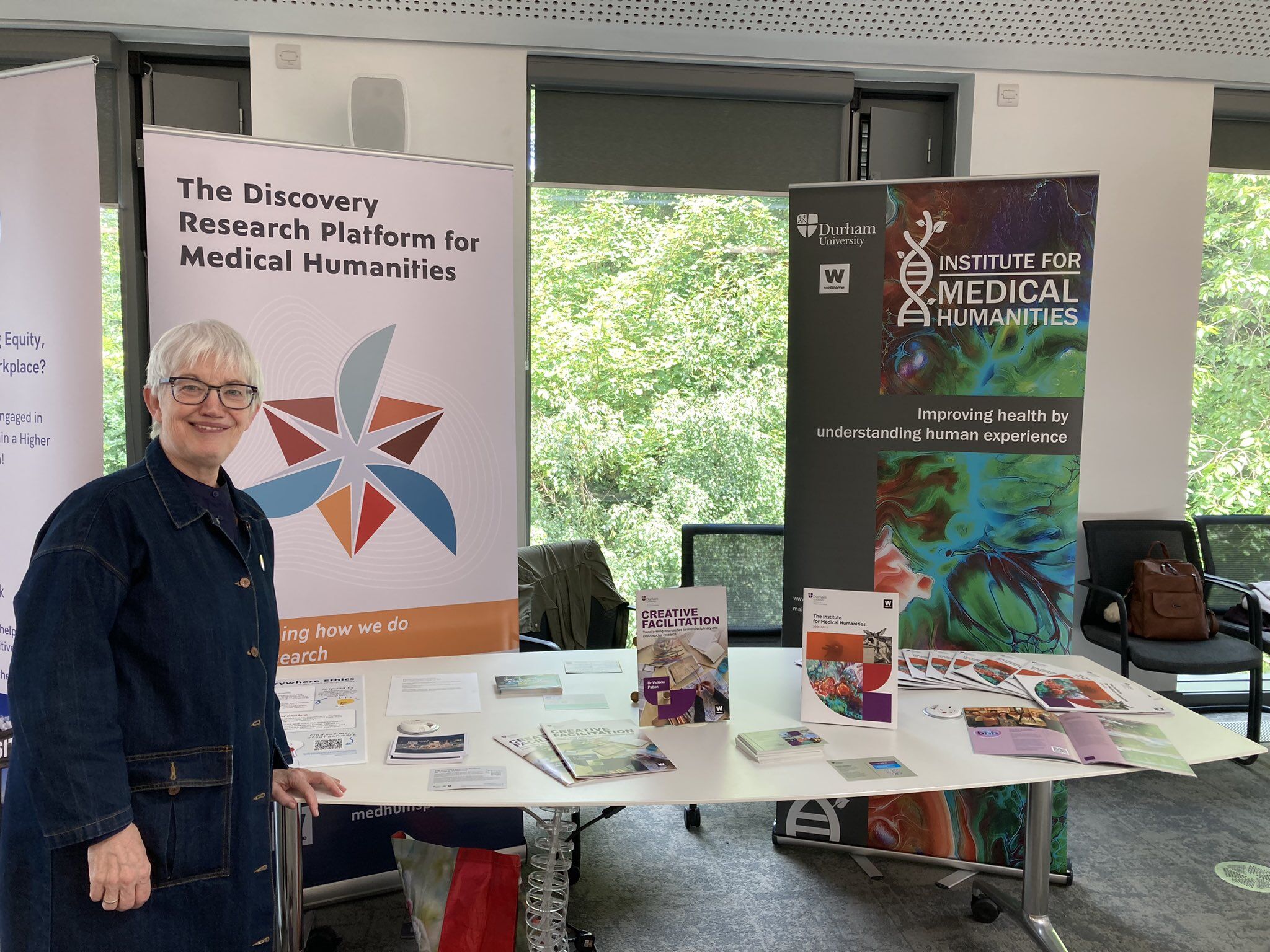 IMH EDI Lead Mary Robson smiles at the camera, standing in front of a stall featuring print materials on IMH practices.