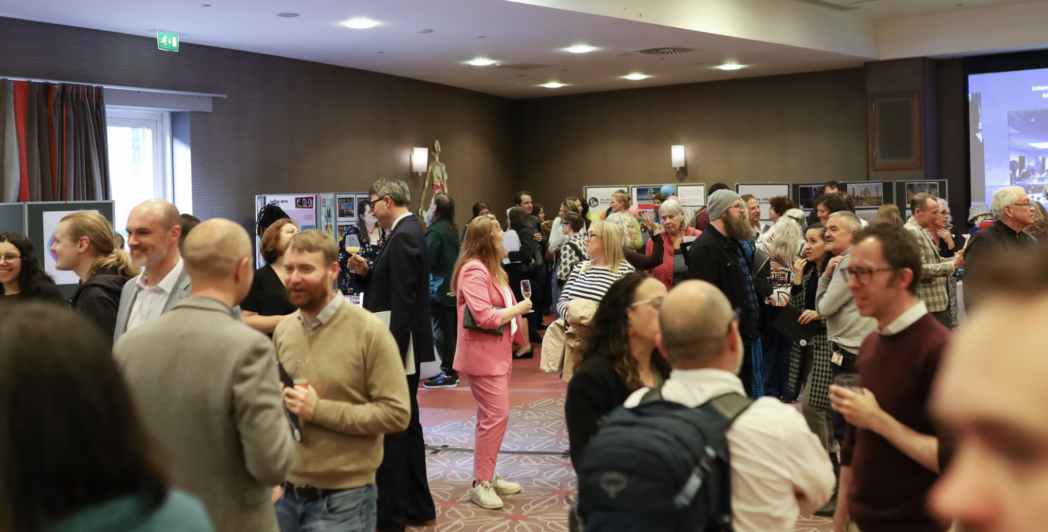 A large room full of people talking and drinking and viewing multiple displays. There are red curtains on the windows and there is a large screen with slides on it