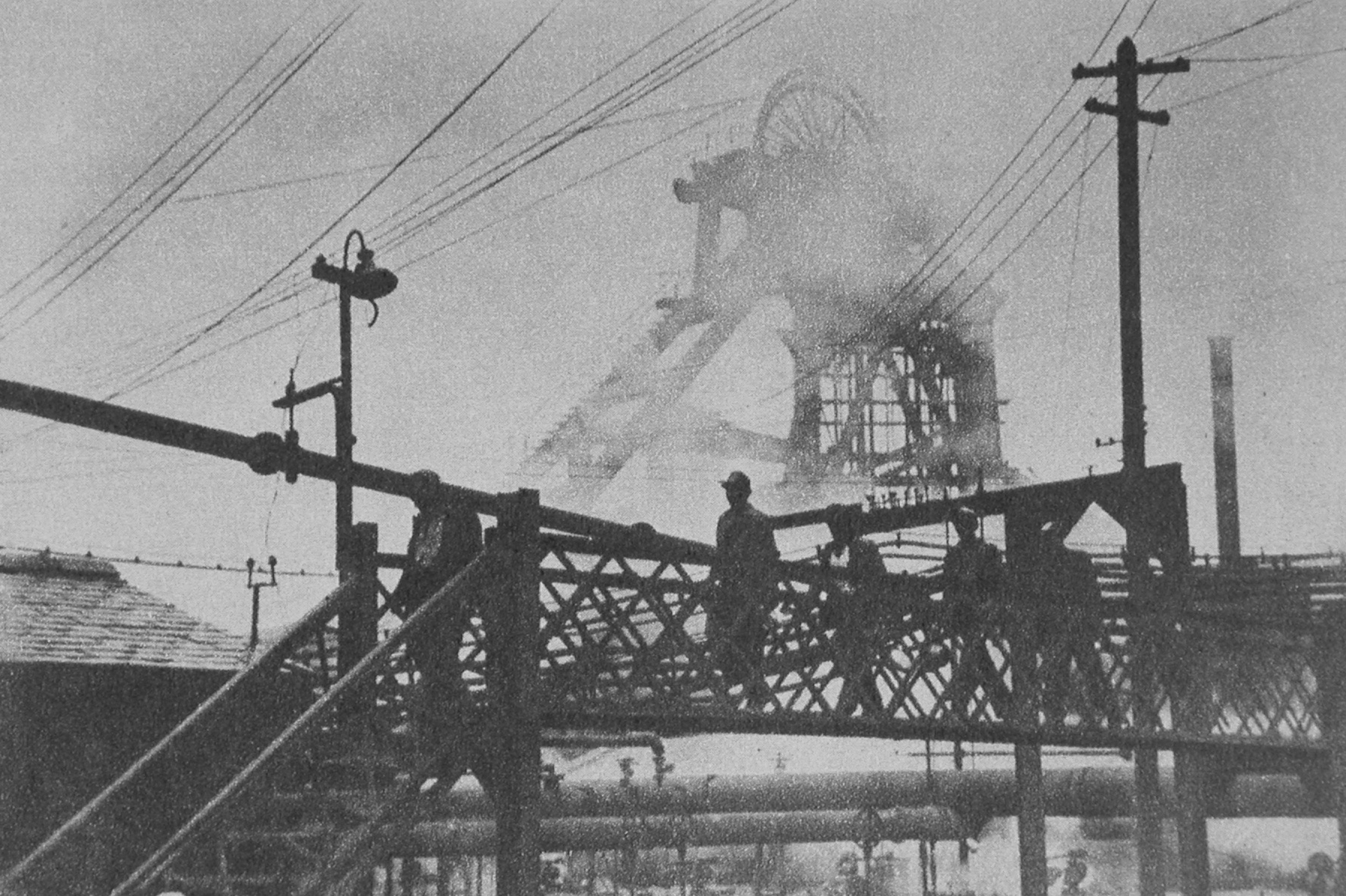 Old black and white photograph of a coal mine. At the forefront of the image is a bridge, across which a group of miners walk.