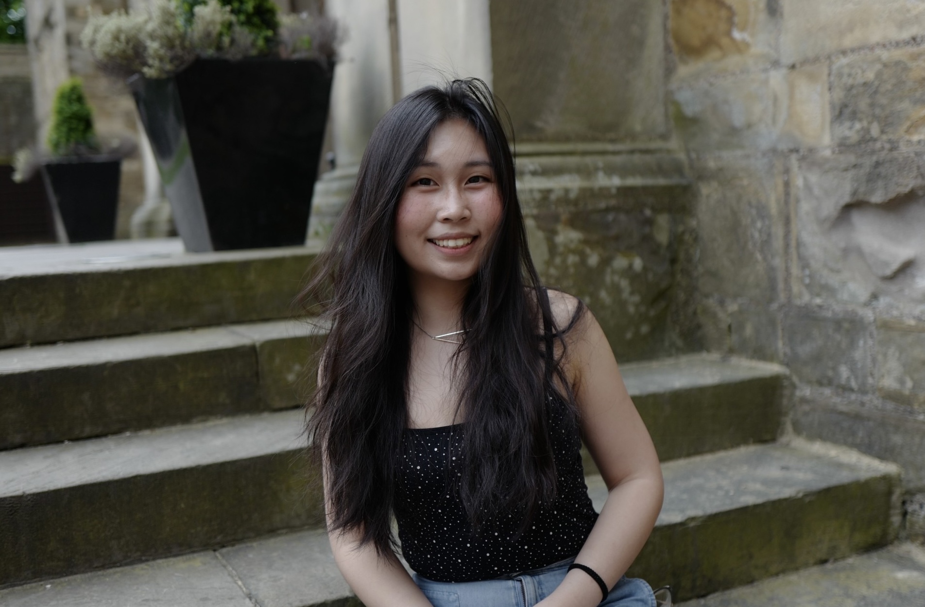 Joyanne Chan, a young woman with black hair, wearing a black top and jeans, sits on a flight of castle steps.