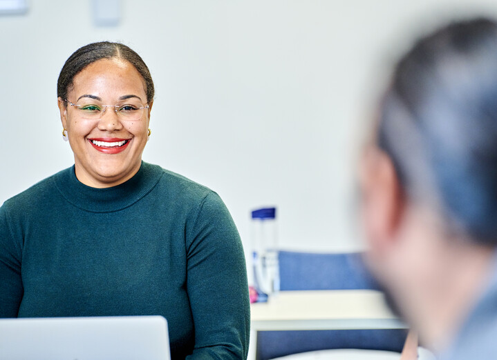 A woman with black hair pulled back off her face and clear framed glasses smiles at colleagues off camera. She wears a dark grey turtle neck jumper and is sitting inside a meeting room. She is an associate professor in the Sociology department