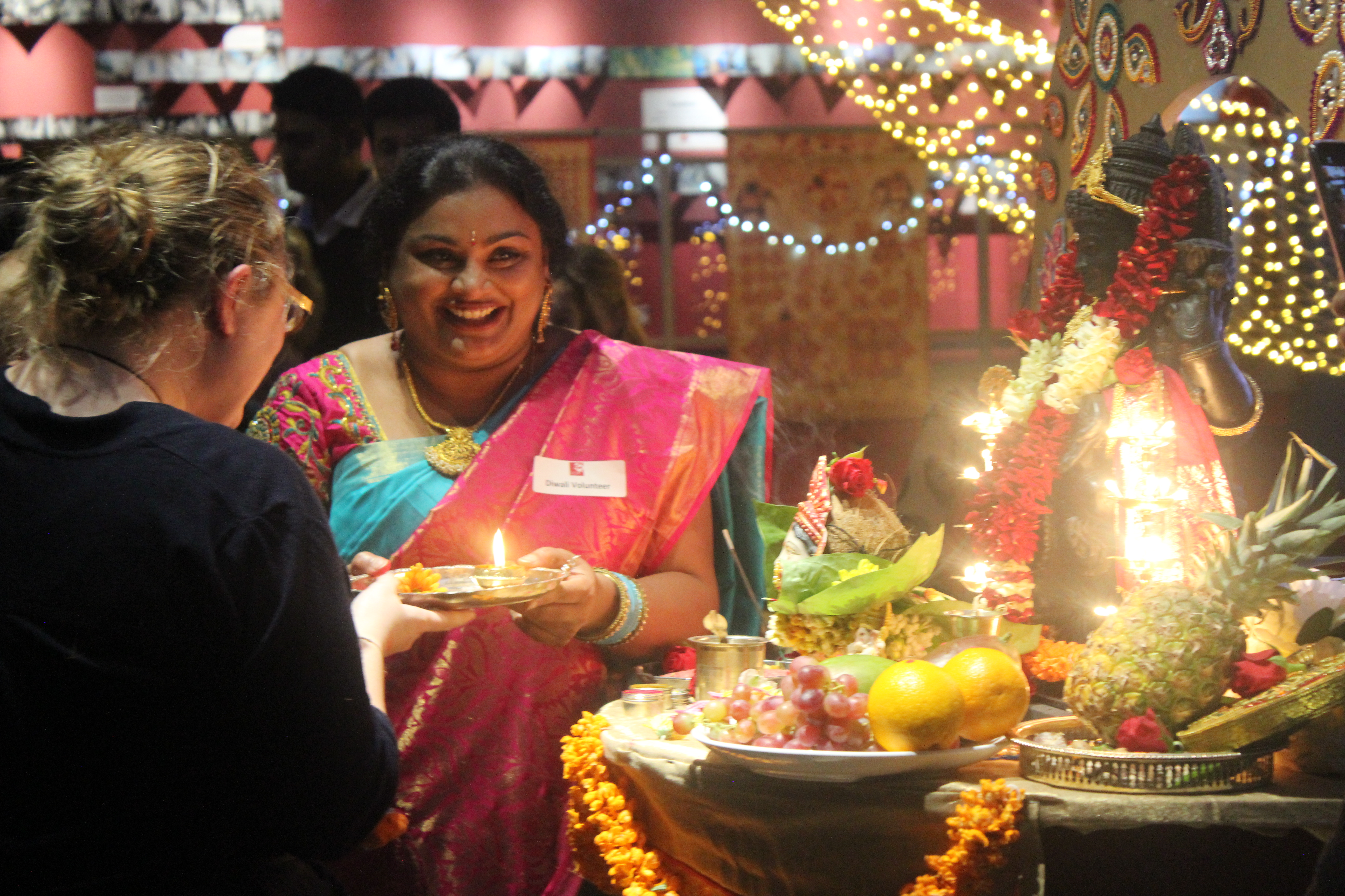 Community volunteer leading prayers to goddess Lakshmi at Diwali celebrations. Goddess Lakshmi statue is lit up with lights and a range of offerings lie in front.