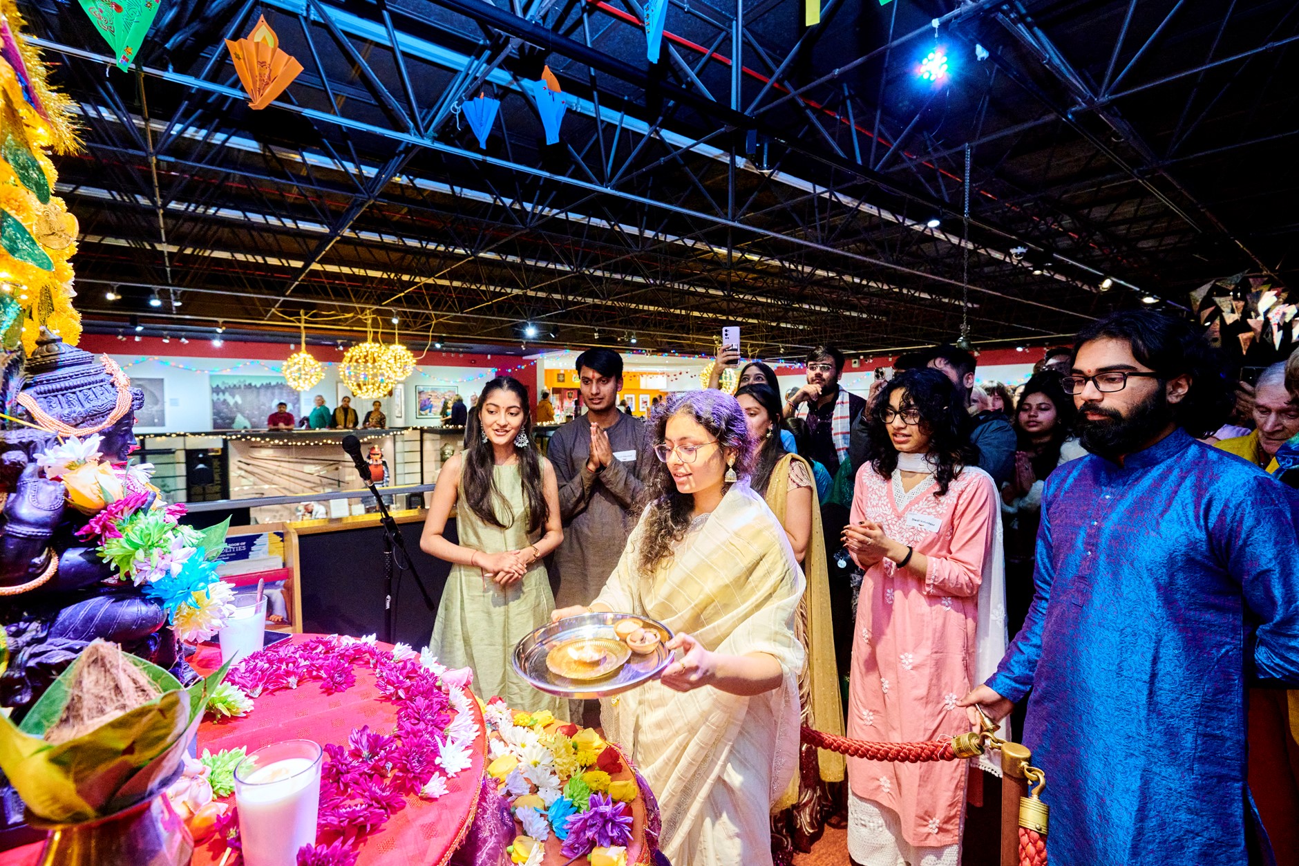 A lady dressed in a sari leads the Diwali Lakshmi Puja ceremony by holding a Puja plate in front of the shrine. The shrine is decorated in flower garlands. Other people stand around the central lady praying with traditional Indian dress.