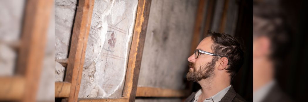 A man with a beard and glasses looks at a fragment of medieval wall painting in a masonry style with a red flower and stem motif