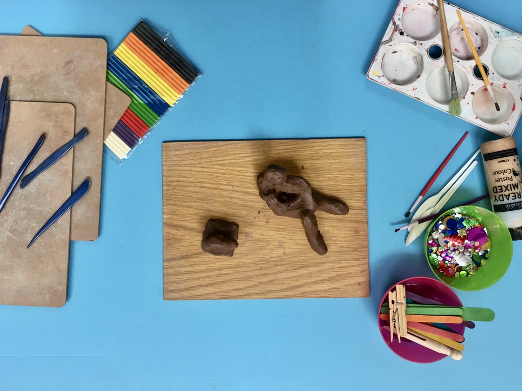 Aerial view of blue table with wooden board in centre. On the board are two pieces of brown clay that have been sculpted in to Henrey Moore style sculptures.